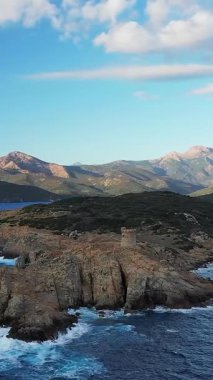 Korsika, Cargese yakınlarındaki engebeli bir kıyı şeridinde mavi denizin üzerindeki kayalık uçurumlar ve dağlık zemin üzerinde tarihi bir Ceneviz kulesi yer alıyor..