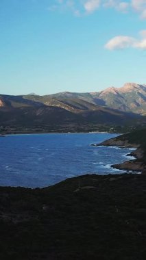 Korsika, Cargese yakınlarındaki engebeli kıyı şeridi ve dağların manzarası, açık mavi bir gökyüzünün altında. Derin mavi deniz yemyeşil tepeler ve kayalık kıyılarla çelişiyor..