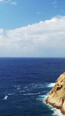 Parçalı bulutlu gökyüzünün altındaki engebeli kayalık bir kıyıya vuran dalgalı açık mavi deniz, Fanale di Petra, Korsika.