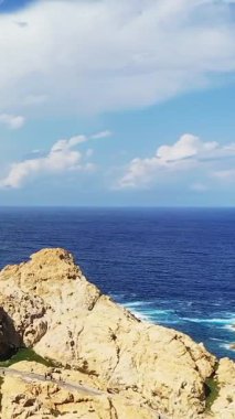 Kayalık uçurumların güneşli manzarası ve Fanale di Petra 'daki derin mavi deniz, canlı bir gökyüzü ve kıyı şeridi üzerinde dağınık bulutlar..