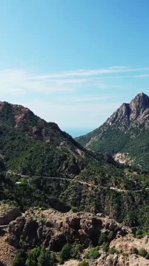 Korsika kırsalındaki engebeli dağların ve dolambaçlı yolların berrak mavi gökyüzünün altında manzarası. Bereketli yeşillik kayalık araziyi kaplıyor. Uzak tepeler ve ufukta deniz manzarası var..