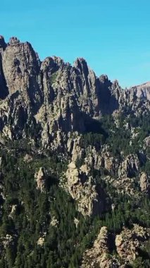 Korsika 'nın Col de Bavella dağlarındaki açık mavi gökyüzünün altında yoğun bir çam ormanının üzerinde dik kayalıklar yükselir..