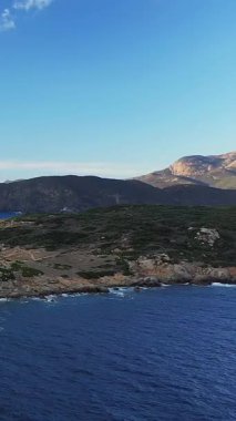 Korsika, Cargese yakınlarında engebeli kıyı şeridi ve arkasında dağlar olan mavi denizin manzarası. Açık gökyüzü ve doğal Akdeniz bitkisi sakin bir atmosfer yaratır..