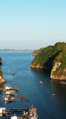 Cat Ba Adası, Vietnam yakınlarındaki yemyeşil kayalıklarla çevrili sakin mavi sularda yüzen teknelerin hava görüntüsü. Yüzen yapılar ve manzaralı kireçtaşı oluşumları berrak gökyüzünün altında görülebilir..