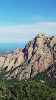 Col de Bavella, Korsika 'da açık mavi bir gökyüzü altında yemyeşil ormanların üzerinde dramatik kayalık dağlar yükselir. Seyahat ve macera kavramları için ideal doğal bir manzara..