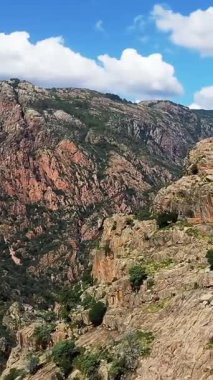 Korsika, Vallee de Porto 'da dağınık bulutlu, parlak mavi bir gökyüzü altında dramatik kayalık ve engebeli dağlar. Yeşil bitki örtüsü ve çarpıcı jeolojik oluşumlarla doğal manzara.