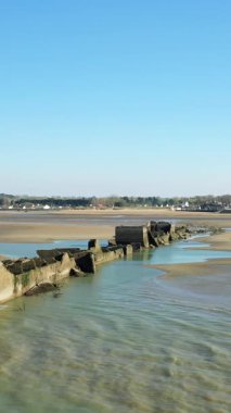 Beton bir yapay liman yapısının kalıntıları, açık mavi gökyüzünün altındaki Asnelles, Normandiya 'daki Gold Beach' teki sığ sulara kadar uzanıyor. Sahne, sakin bir kıyı manzarası yakalar ve gelgitte tarihi kalıntılar görünür olur..