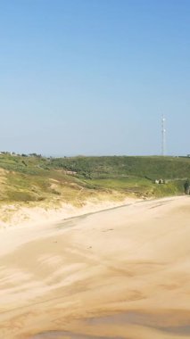 Fransa 'nın Cap de Carteret şehrinde açık mavi gökyüzünün altında çimenli kayalıklarla çevrili geniş kumlu bir sahil. Ufukta uzun bir iletişim kulesi duruyor ve doğal manzaraya modern bir dokunuş ekliyor..