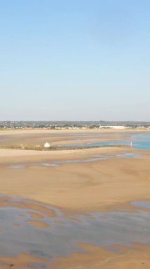 Plage de la Potiniere 'in açık mavi gökyüzünün altında altın kumu ve uzak bir köy manzarası. Sakin kıyı manzarası doğa ve seyahat kavramları için idealdir..