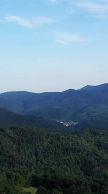Açık mavi gökyüzünün altında Fransa 'nın Vosges bölgesindeki sık orman ve kayan tepelerin manzarası. Vadideki küçük bir köy yemyeşil alanlarla çevrili..