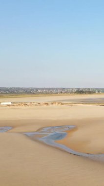 Plage de la Potiniere 'de açık mavi gökyüzünün altında sığ gelgit havuzları olan geniş kumlu sahil. Ufukta görünen uzak kasaba ve kıyı şeridi.