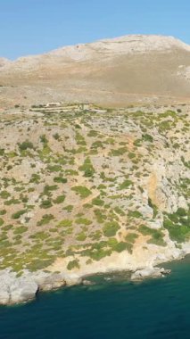 Girit 'teki Preveli Sahili yakınlarında engebeli bir yamacın, seyrek bitki örtüsünün turkuaz denizle buluşması. Parlak güneş ışığı kıyı manzarasının doğal dokusunu vurgular.