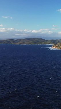 Dalgaların kayalık bir kıyı şeridine çarptığı engin mavi deniz ve açık gökyüzünün altındaki uzak yeşil tepeler, Les Iles Sanguinaires, Korsika.