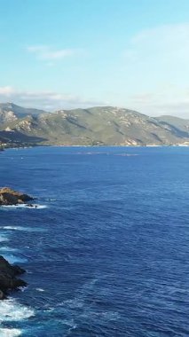 Les Iles Sanguinaires, Korsika yakınlarındaki engebeli kıyı şeridi ve derin mavi suların manzarası, berrak bir gökyüzü altında uzak dağlar..