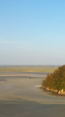 Fransa 'nın Le Mont-Saint-Michel yakınlarındaki açık mavi gökyüzünün altında kayalık bir çıkıntı ve bitki örtüsü olan kumlu gelgit dairesinin geniş manzarası..