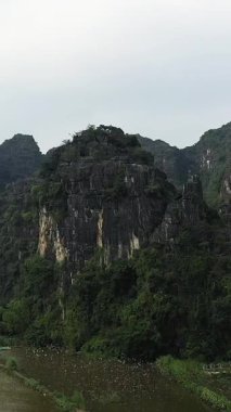 Yoğun yeşil bitki örtüsüyle kaplı dramatik kireçtaşı uçurumları Ninh Binh, Vietnam 'da sakin bir kırsal alanın üzerinde yükseliyor. Sahne, doğal güzellikleri ve eşsiz jeolojik oluşumları yakalıyor..