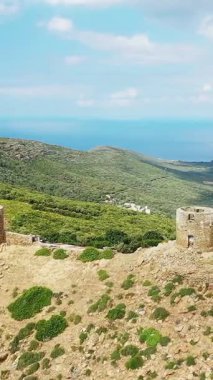 Korsika 'daki Col de la Serra' daki yemyeşil tepelere ve uzak denize bakan eski bir taş kulenin havadan görünüşü. Parlak mavi gökyüzünün altındaki engebeli manzara.