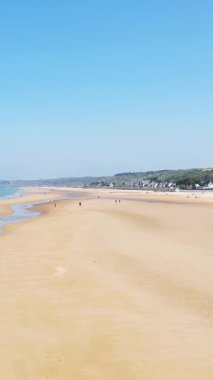 Omaha Sahili 'nin geniş manzarası. Altın kumlar, birkaç uzak insan ve açık mavi gökyüzü altında Normandiya kıyısı boyunca küçük evler..
