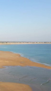 Geniş kumlu plaj ve sakin mavi deniz Plage de la Potiniere 'de berrak bir gökyüzü altında, kıyı boyunca uzak evler. Huzurlu kıyı manzarası seyahat ve rahatlama temaları için ideal.