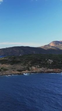 Korsika, Cargese yakınlarındaki engebeli kıyı şeridi ve yuvarlanan tepelerin berrak mavi gökyüzünün altında manzarası. Manzarada Akdeniz kıyısındaki kayalık kıyılar ve yemyeşil alanlar yer alıyor..