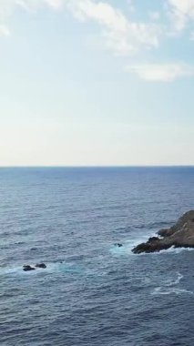 Dalgalar, Korsika, Cargese yakınlarında parlak bir gökyüzünün altında engebeli bir kayalık kıyıya çarpar. Berrak mavi su ve dağınık bulutlarla sakin bir deniz manzarası..