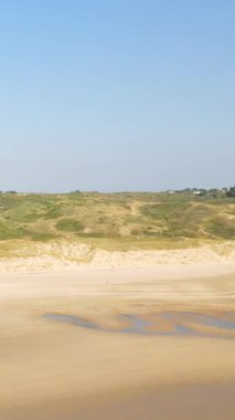 Plage de la Vieille Eglise 'de yumuşak kum tepeleri ve açık mavi gökyüzü olan geniş kumlu bir sahil. Doğal yeşillik ve gelgit havuzlarıyla huzurlu kıyı manzarası.