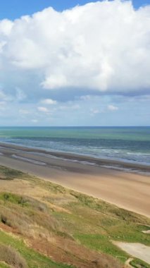 Fransa, Normandiya 'daki Omaha Sahili' nin geniş kumlu kıyı şeridinde hafif dalgalar ve dramatik bulutlar var. Çimenli kum tepeleri ve açık denizli huzurlu kıyı manzarası.