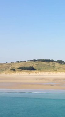 Plage de la Vieille Eglise 'de açık mavi gökyüzünün altında yumuşak dalgalar ve çimenli kum tepeleri olan geniş kumlu bir sahil. Huzurlu kıyı manzarası, doğa ve seyahat kavramları için ideal..