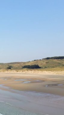 Plage de la Vieille Eglise 'de açık mavi gökyüzünün altında yumuşak dalgalar ve çimenli kum tepeleri olan geniş kumlu bir sahil. Huzurlu kıyı manzarası doğa ve seyahat kavramları için ideal..