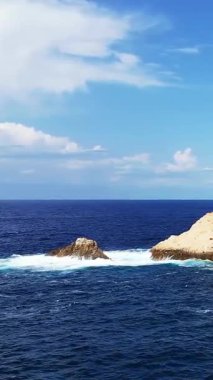 Açık mavi okyanus dalgaları, Korsika 'daki Fanale di Petra yakınlarında parlak bir gökyüzü altında kayalık çıkıntılara çarpar. Dramatik kıyı manzaralı sakin bir deniz manzarası.