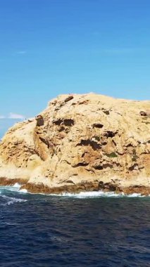 Sardinya, Fanale di Petra 'da açık mavi bir gökyüzünün altında engebeli bir kayaya dalgalar çarpar. Dramatik kıyı şeridinde desenli taş ve canlı deniz renkleri yer alıyor..