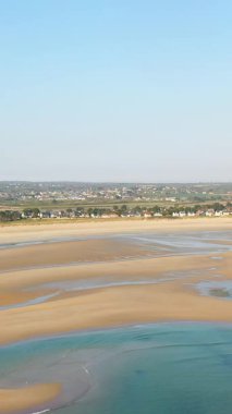 Geniş kumlu plaj gelgit havuzları ve açık mavi gökyüzü altında uzak evler Grande Plage de Barneville, Fransa.