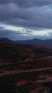 Yunanistan 'ın Kardamyli kenti yakınlarındaki kasvetli bir akşam gökyüzünün altındaki engebeli kıyı dağlarının manzarası. Dramatik bulutlar ve dolambaçlı yollar çarpıcı bir doğal manzara yaratır..