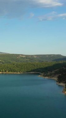 Yoğun yeşil ormanlarla çevrili sakin bir göl ve berrak mavi gökyüzünün altındaki yumuşak tepelerin huzurlu manzarası. Doğal manzara sakinlik ve açık hava eğlencesi kavramları için idealdir..