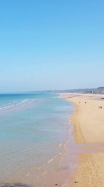 Fransa, Normandiya 'daki Omaha Sahili' nin geniş manzarası, yumuşak dalgalar, altın kum ve açık mavi gökyüzü. Birkaç kişi huzurlu kıyı şeridinde geziniyor..