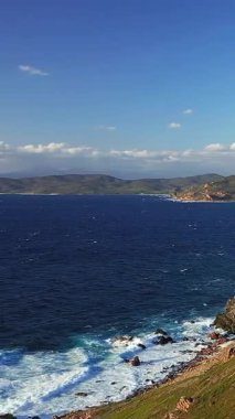 Açık bir gökyüzünün altında, Les Iles Sanguinaires, Korsika 'da kayalık kıyı ve derin mavi deniz manzarası. Dalgalar ufuktaki uzak tepelerle engebeli kıyı şeridine çarpar..