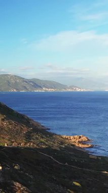 Engebeli kıyı şeridinin manzarası ve Les Iles Sanguinaires, Korsika yakınlarındaki berrak mavi sular, parlak bir gökyüzü altında uzak dağlar..
