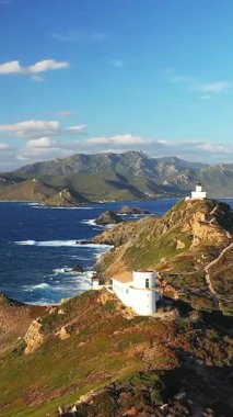 Les Iles Sanguinaires 'in engebeli sahil şeridine tünemiş beyaz bir deniz fenerinin manzarası, dramatik dağlar ve parlak bir gökyüzünün altında mavi deniz..