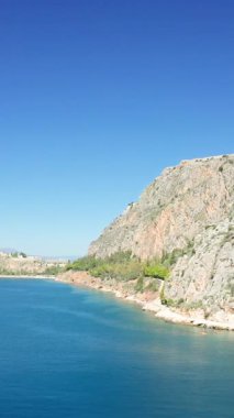 Clear blue water meets a rugged rocky coastline under a bright sky in Nafplio, Greece. Natural landscape with steep cliffs and lush greenery along the shore.