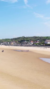 Fransa, Normandiya 'daki Omaha Sahili' nin geniş manzarası kumlu bir kıyı şeridi, uzak insanlar ve mavi gökyüzünün altındaki liman boyunca birden fazla bayrak..