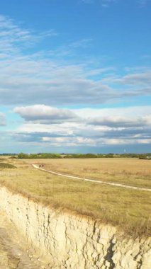 Dramatik bir uçurum kenarı, açık otlak alanları ve dağınık bulutlu geniş mavi bir gökyüzü içeren geniş kırsal bir manzara. Huzurlu kırsal yol huzur hissi katıyor..