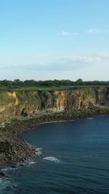 Fransa 'nın Pointe du Hoc kentindeki engebeli uçurumların ve kayalık kıyı şeridinin hava manzarası açık mavi bir gökyüzünün altında. Dramatik manzarada sarp kayalıklar, yeşil çimenler ve kayalara çarpan dalgalar var..