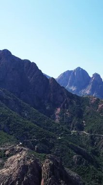 Korsika Vallee de Porto 'daki engebeli dağların ve dolambaçlı yolun manzarası, berrak mavi bir gökyüzünün altında. Yeşiller yemyeşil yamaçlar ve dramatik kayalık tepeler manzaraya hakim..