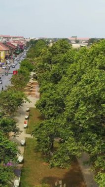 Battambang, Kamboçya 'nın merkezindeki işlek bir caddeye komşu ağaçlar ve banklarla kaplı yeşil bir parkın yüksek açılı görüntüsü..