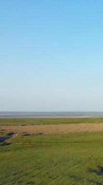 Fransa 'nın Le Mont-Saint-Michel yakınlarındaki açık mavi gökyüzünün altında çimenli tarlaların ve gelgit düzlüklerinin geniş bir görüntüsü. Doğal dokuları ve yumuşak ışığı olan sakin kırsal manzara..