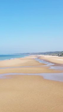 Geniş kumlu kıyı şeridinin ve Fransa 'nın Normandiya kentindeki Omaha Sahili' ndeki açık mavi gökyüzünün altında hafif dalgaların havadan görünüşü. Sahilde yürüyen çok az insan, uçsuz bucaksız ve sakin manzarayı vurguluyor..