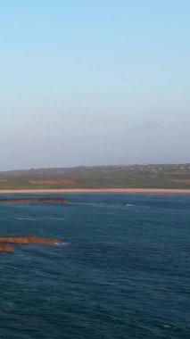 Cap de la Hague, Normandiya, Fransa 'daki engebeli kıyı şeridi ve mavi okyanus manzarası açık bir gökyüzünün altında. Sahnede kayalık çıkıntılar, yuvarlanan alanlar ve sakin bir deniz manzarası yer alıyor..