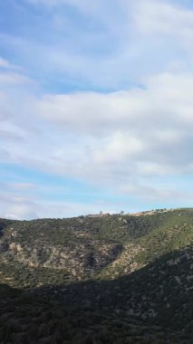 Parçalı bulutlu gökyüzünün altındaki yeşil tepelerin geniş bir görüntüsü Hermione yakınlarındaki doğal manzarayı yakalıyor. Sahnede engebeli arazi ve ufuktaki uzak ağaçlar yer alıyor..