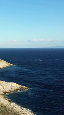 Engebeli kayalık bir kıyı şeridinin açık bir gökyüzünün altında derin mavi denizle buluşmasını geniş açıdan görmek. Doğal sahil güzelliği ile sakin deniz manzarası.