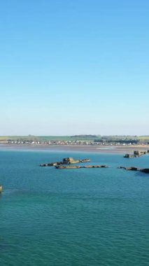 Asnelles, Normandiya açıklarında, parlak bir gökyüzünün altında yapay bir limanın kalıntıları olan berrak mavi sular. Arka planda kıyı şeridi ve uzak köy görülüyor..